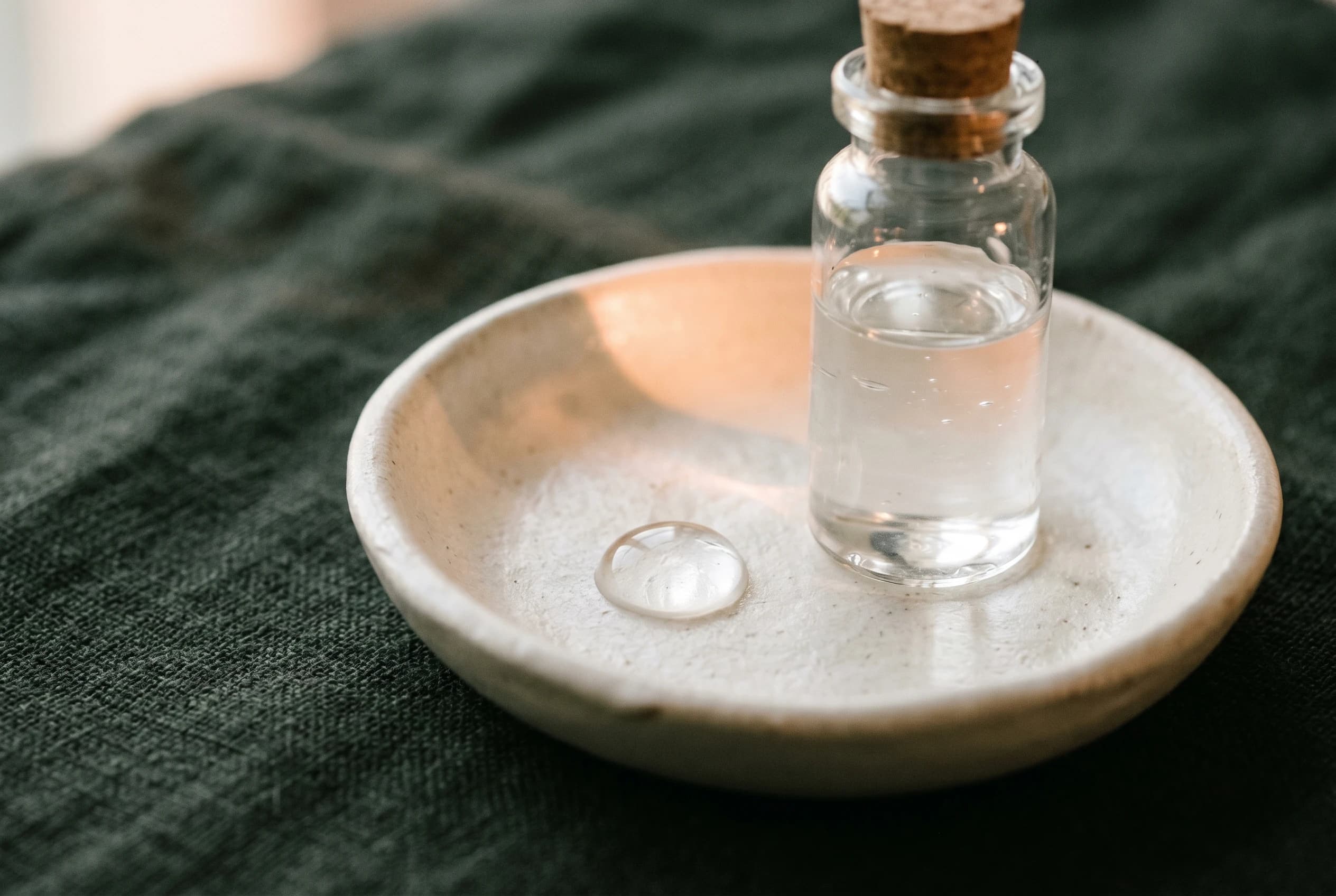 Klares Hyaluron-Gel in einem Fläschchen und ein Tropfen in einer bone-cream Keramikschale auf charcoal-grünem Leinen — Symbol der reversiblen Hyaluronsäure